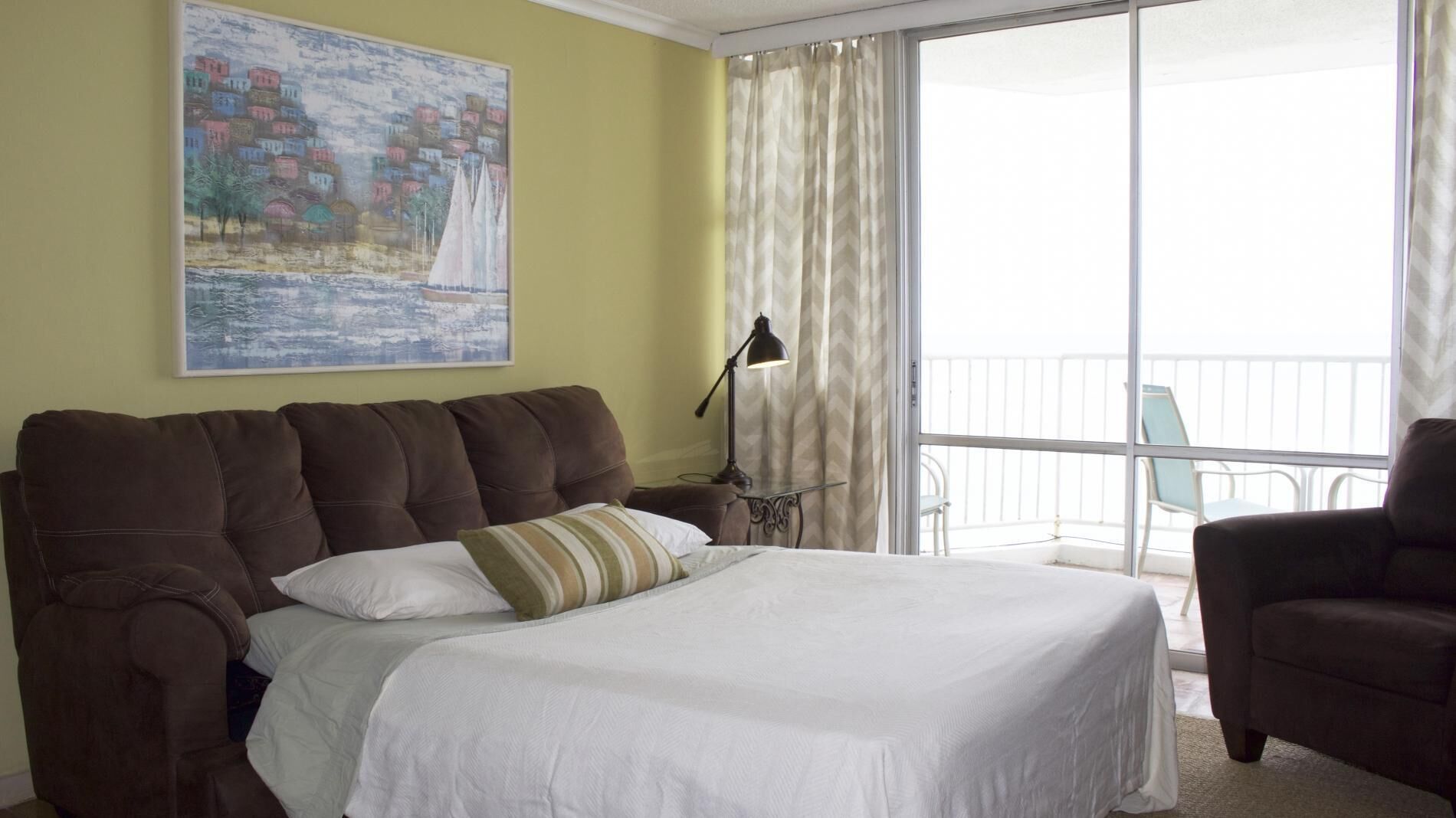 A hotel room with a large bed, a nightstand with a lamp, a balcony with a chair, and a painting on the wall.