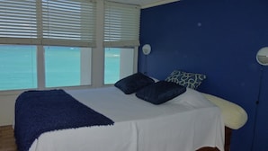 A bedroom with a bed, two pillows, a blue wall, and a window with blinds. - El Capitan (Carolina)