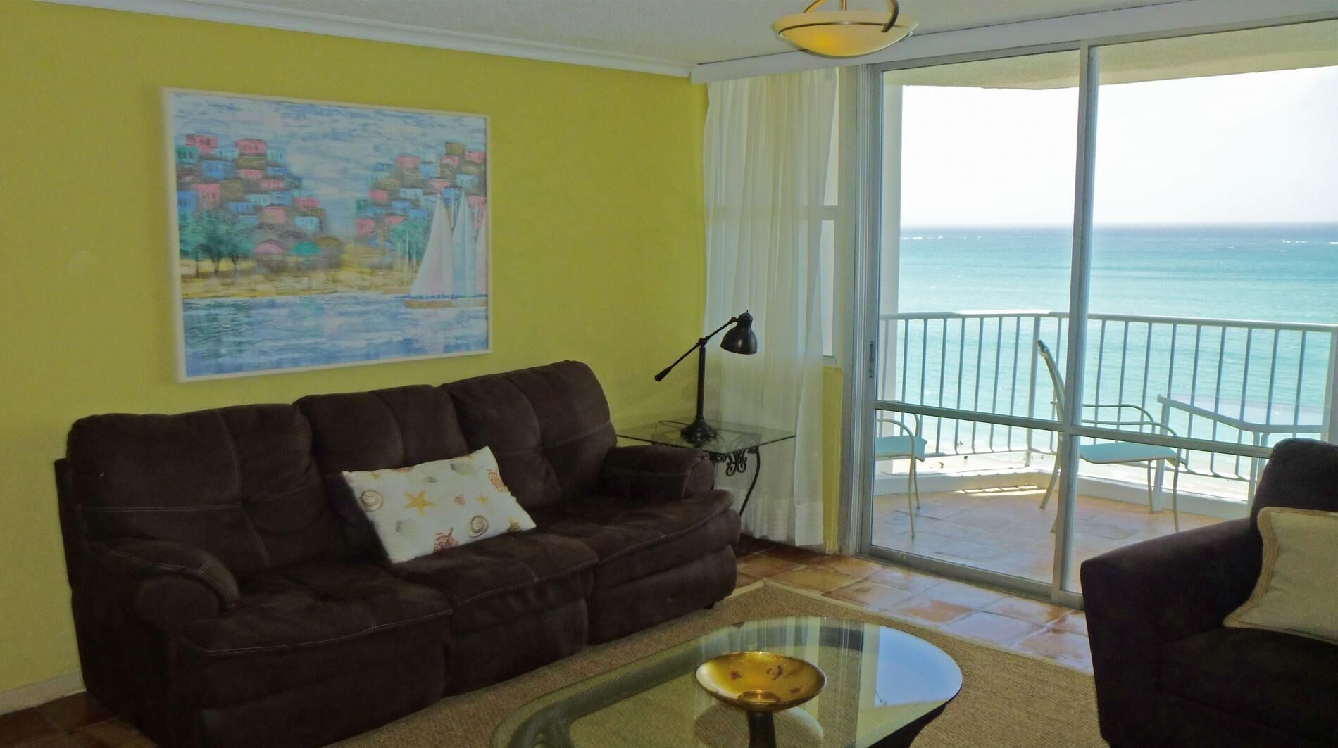 Een woonkamer met een bruine bank, een glazen salontafel, een schilderij aan de muur en een balkon met uitzicht op zee.