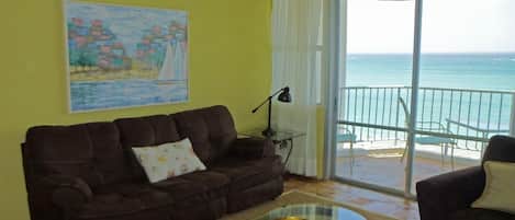 Een woonkamer met een bruine bank, een glazen salontafel, een schilderij aan de muur en een balkon met uitzicht op zee.