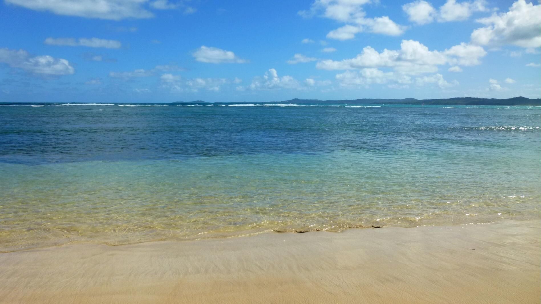 Une plage avec une eau claire et peu profonde, et une chaîne de montagnes qui se dessine à l'horizon.