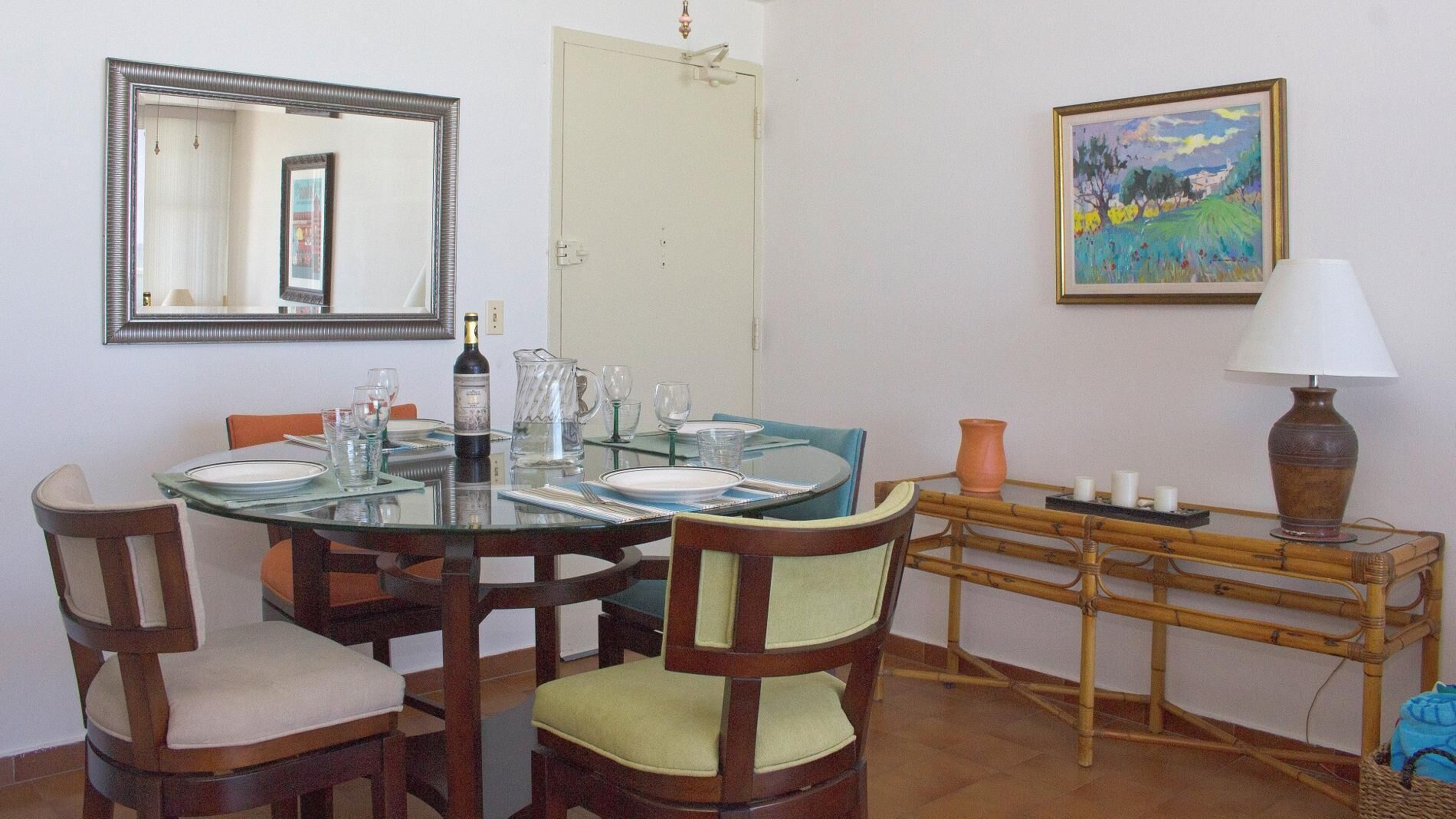 A dining area with a glass table, wooden chairs, a mirror, a painting, and a lamp.