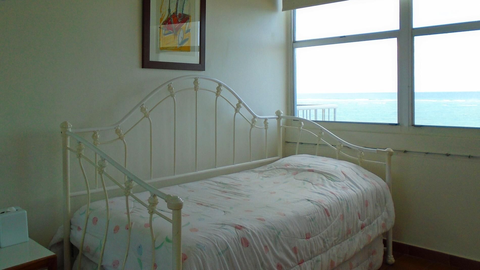 Una habitación pequeña con cama individual, estructura metálica blanca, ropa de cama floral y ventana con vista al mar.