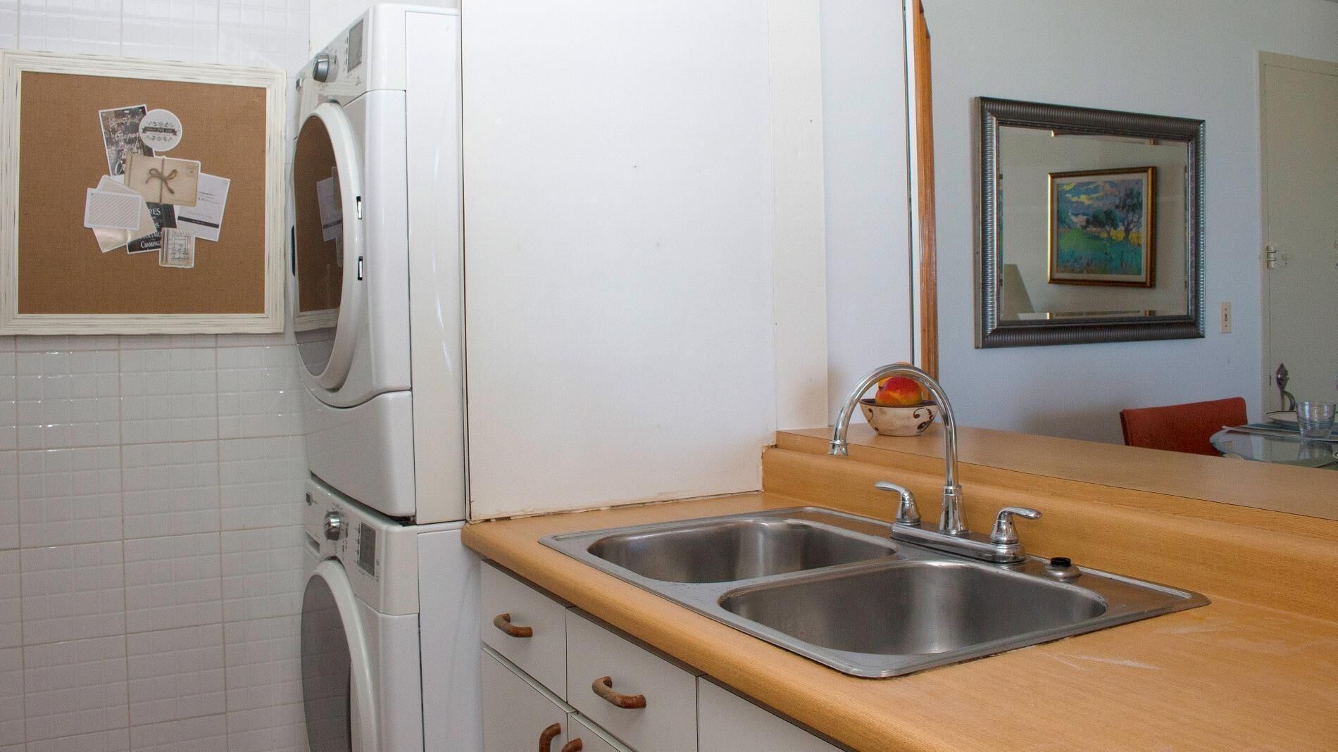 A kitchen with a double sink, a washing machine, and a corkboard with photos and notes.
