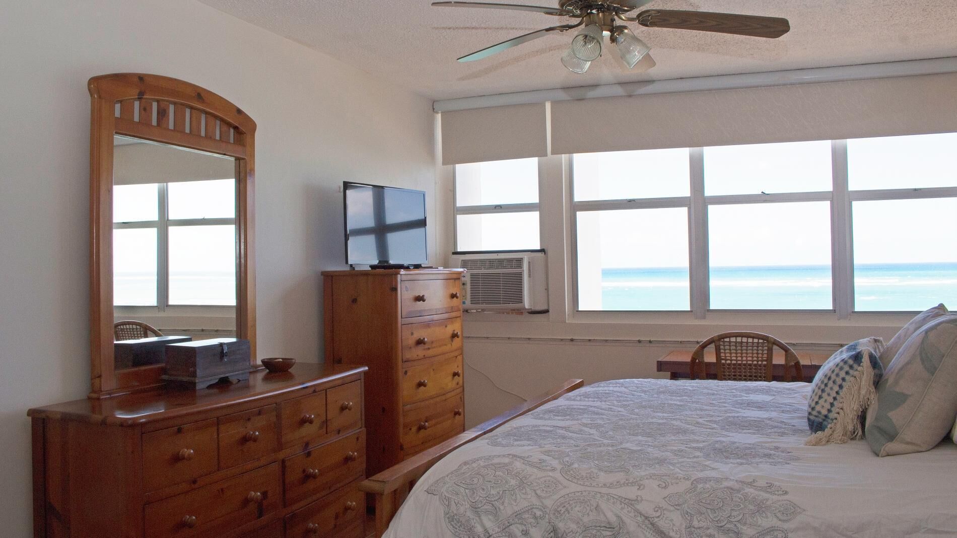 A bedroom with a bed, dresser, television, and a window with a view of the ocean.