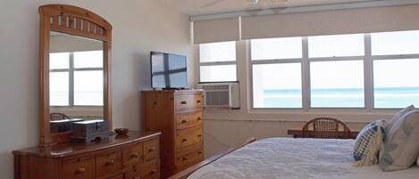 A bedroom with a bed, dresser, television, and a window with a view of the ocean.