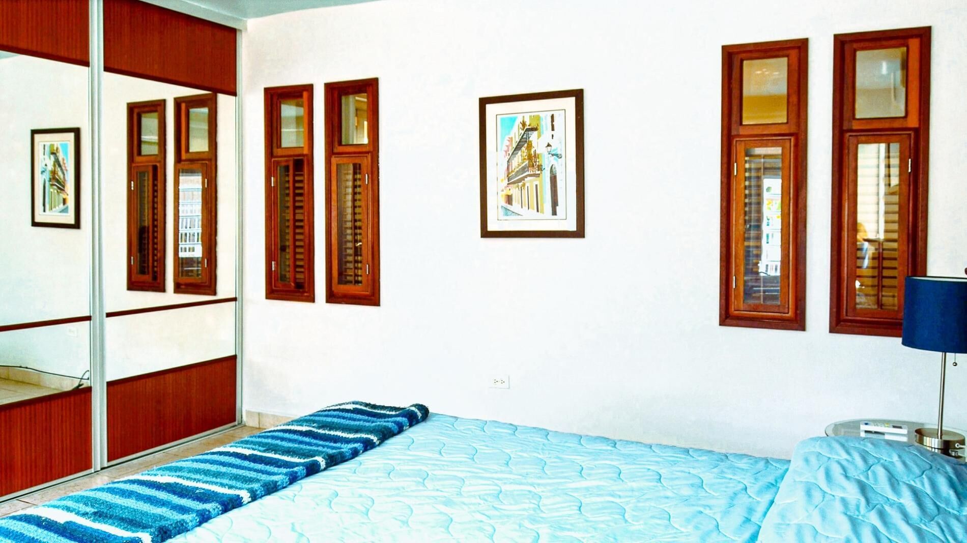 A bedroom with a bed, a blue striped rug, wooden windows, and a framed picture on the wall.