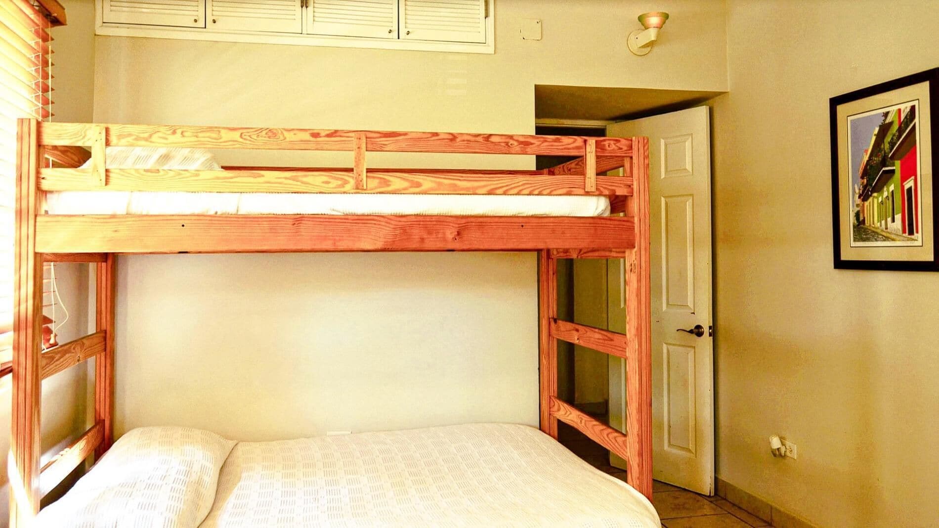 Un lit superposé avec une structure en bois et un matelas blanc. Il y a une porte sur la droite et un tableau encadré au mur.