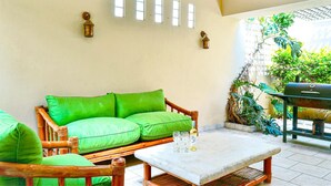 A room with a green cushioned sofa, a small table with glasses and a bottle, and a grill outside. - Villa Serena (San Juan)