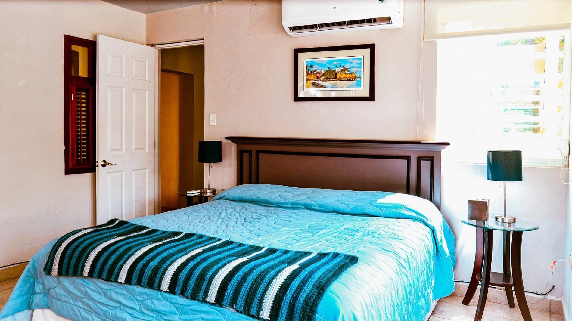 A bedroom with a bed, a nightstand, a lamp, a framed picture on the wall, and an air conditioning unit.