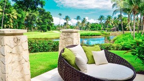 A wicker lounge chair with cushions, set on a tiled patio with a view of a lush garden and a pond. - Villa Margarita (Río Grande)