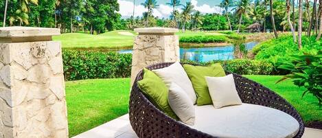 A wicker lounge chair with cushions, set on a tiled patio with a view of a lush garden and a pond.