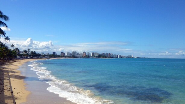 A beach with clear blue water, a sandy shore, and a city skyline in the background. - Villa Verde (San Juan)