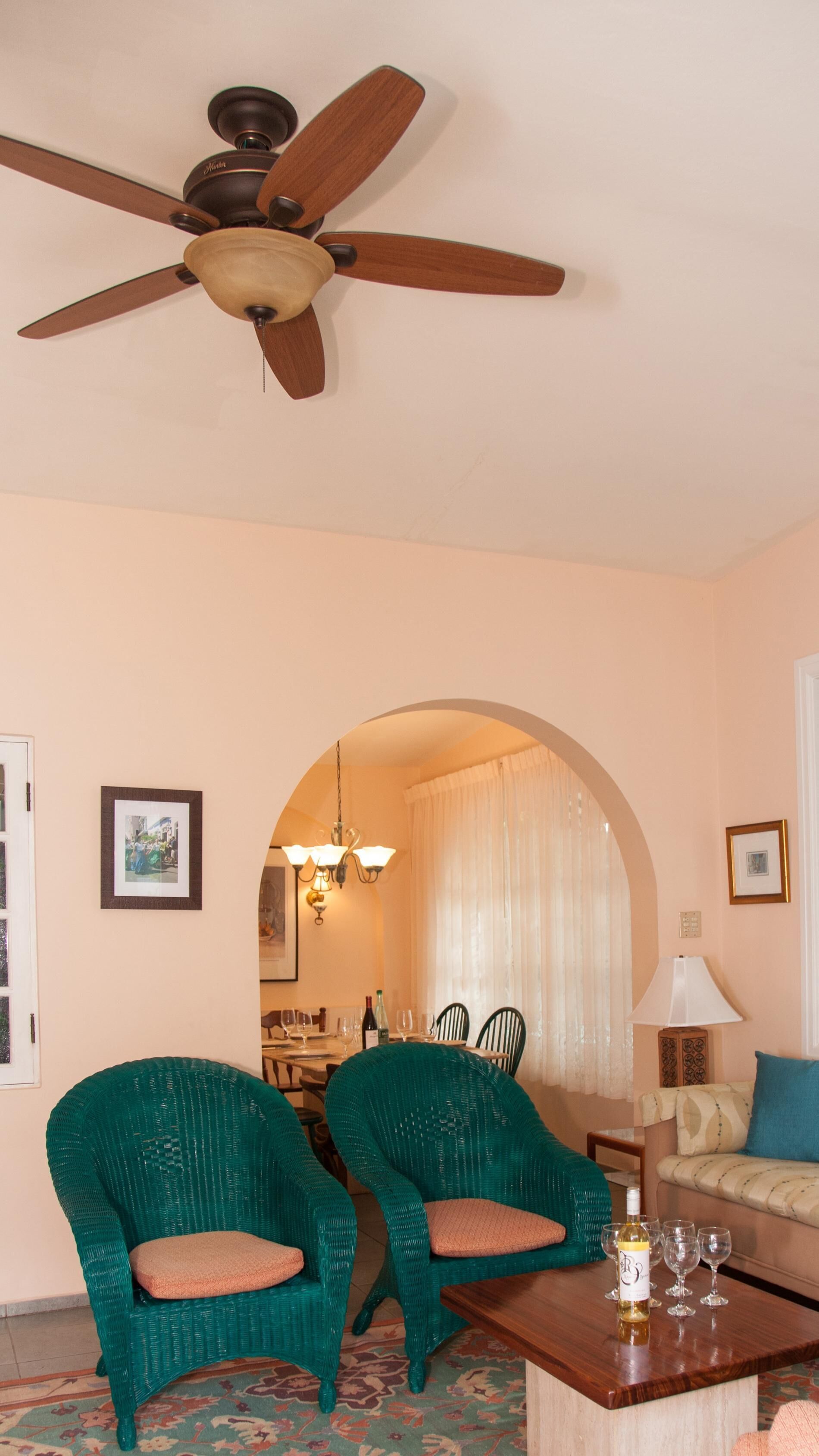 A living room with two green wicker chairs, a wooden coffee table with wine glasses, a ceiling fan, and a doorway leading to another room.