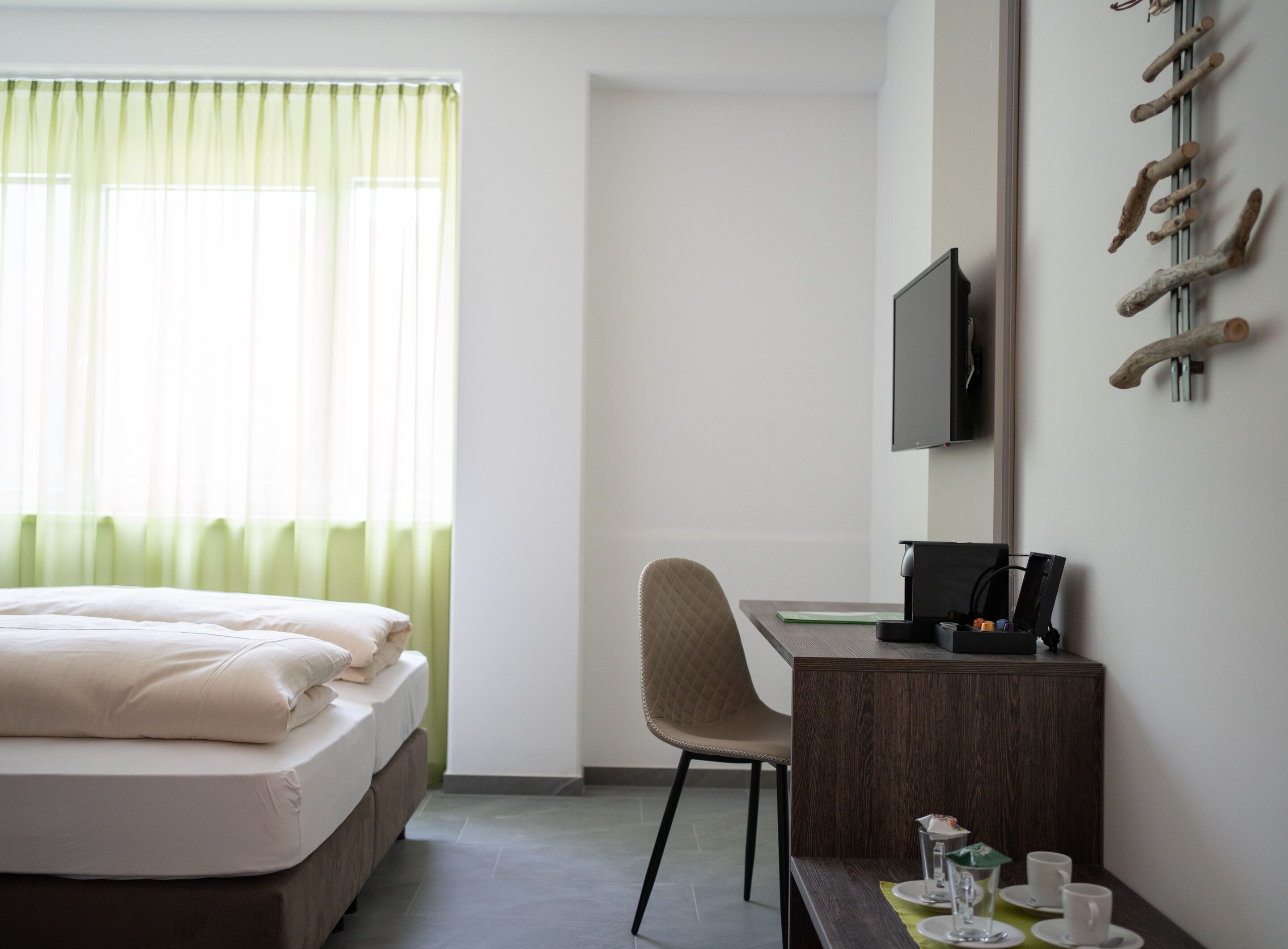 A hotel room with a bed, a desk, a chair, a TV, and a wall-mounted coat rack.