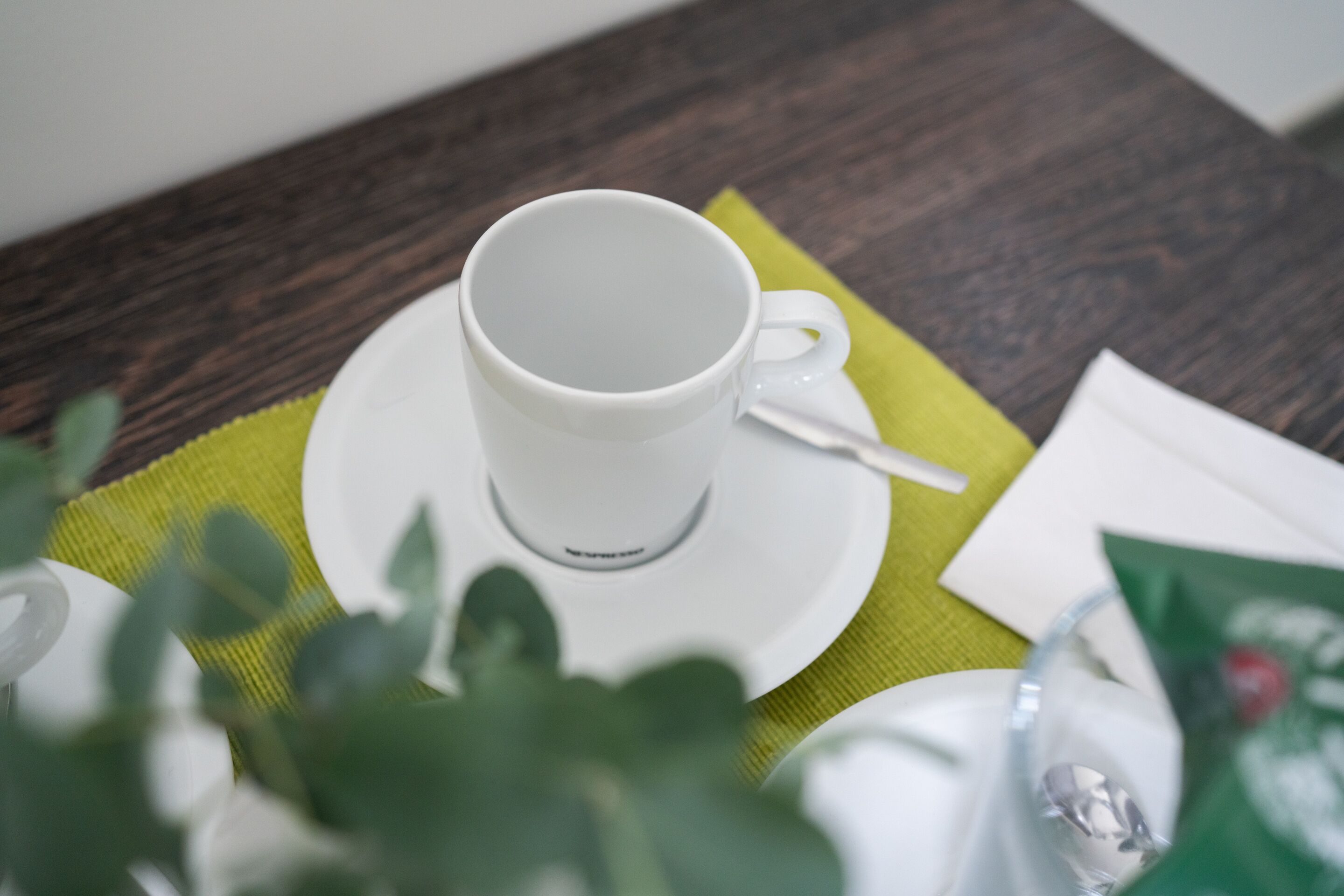 Una taza blanca sobre un platillo con una cuchara, colocada sobre un mantel individual verde en una mesa de madera.