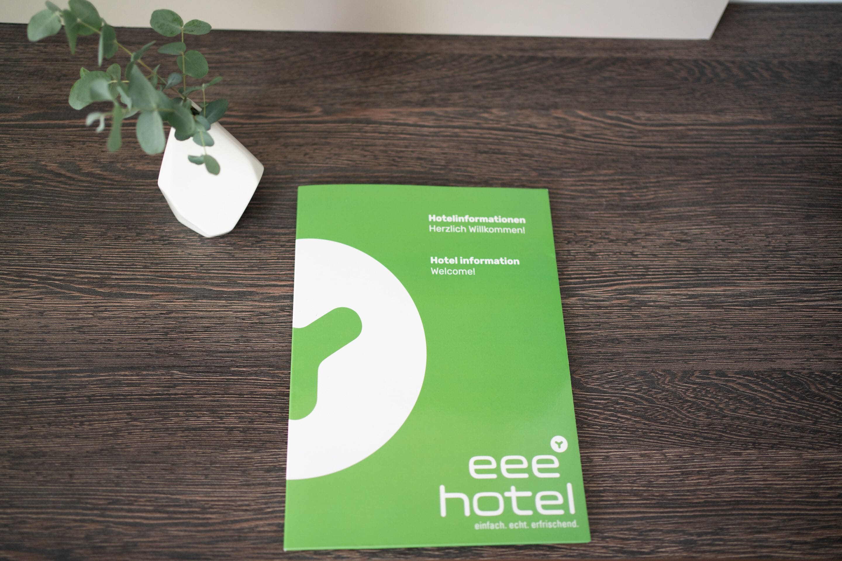 A green booklet titled 'eee hotel' with text in German and English, placed on a wooden surface next to a white vase with green leaves.