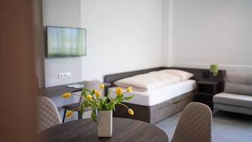 Ein modernes Wohnzimmer mit Esstisch, einer Couch und einem Bett. Ein Fernseher ist an der Wand befestigt und auf dem Tisch steht eine Vase mit Blumen.