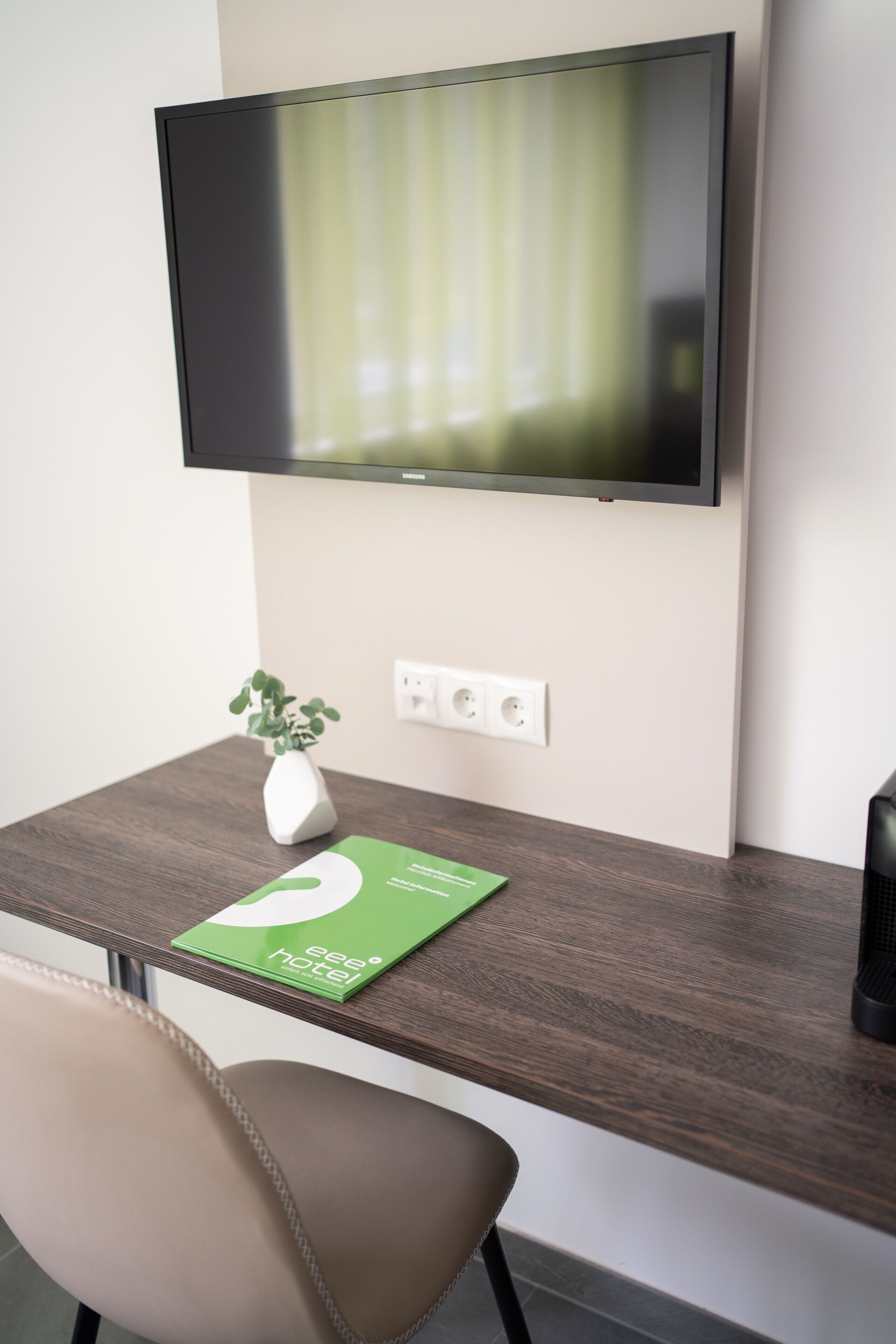 Une chambre d’hôtel moderne équipée d’un téléviseur à écran plat fixé au mur, d’un bureau en bois avec un petit carnet vert, d’une petite plante en pot et d’une chaise beige.