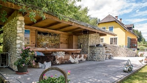 A stone and wood covered outdoor seating area with a stone fireplace, potted plants, and a rustic bench. - Tameggerhof (Gurk)