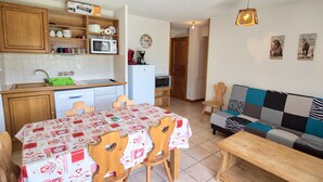 A compact kitchen with wooden cabinets, a microwave, and a refrigerator. There is a dining table with chairs and a patterned tablecloth. A sofa and a bench are in the living area. - Ski-in/ski-out apartment with terrace, pet-friendly, Flumet (Flumet)