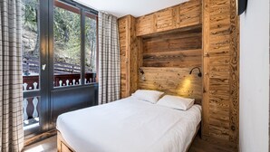 A bedroom with a large bed, wooden panel walls, a window with curtains, and a view of trees. - Renovated Apartment · Near Center · Ski-In/Ski-Out · South-Facing Balcony (Val-d'Isère)