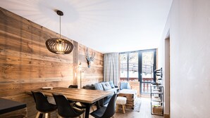 A dining area with a wooden table and chairs, a hanging light fixture, and a large window with a view of the outdoors. - Renovated Apartment · Near Center · Ski-In/Ski-Out · South-Facing Balcony (Val-d'Isère)