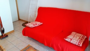 Ein rotes Sofa mit gemusterten Kissen, eine weiße Klapptreppe und ein Staubsauger in einem Raum mit weißen Wänden und Fliesenboden.