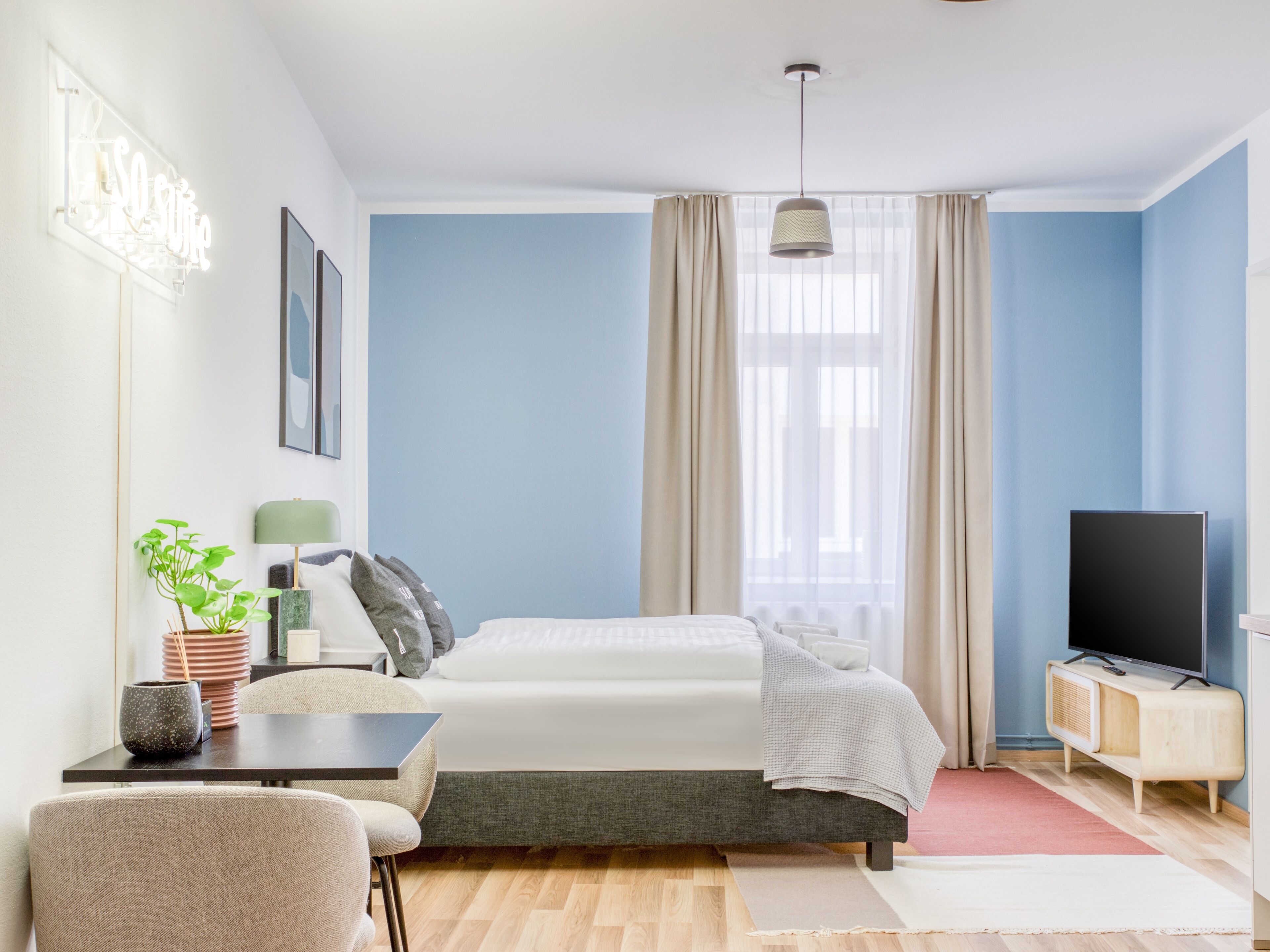 Quarto moderno com cama, escrivaninha, cadeira, televisão e janela com cortinas.