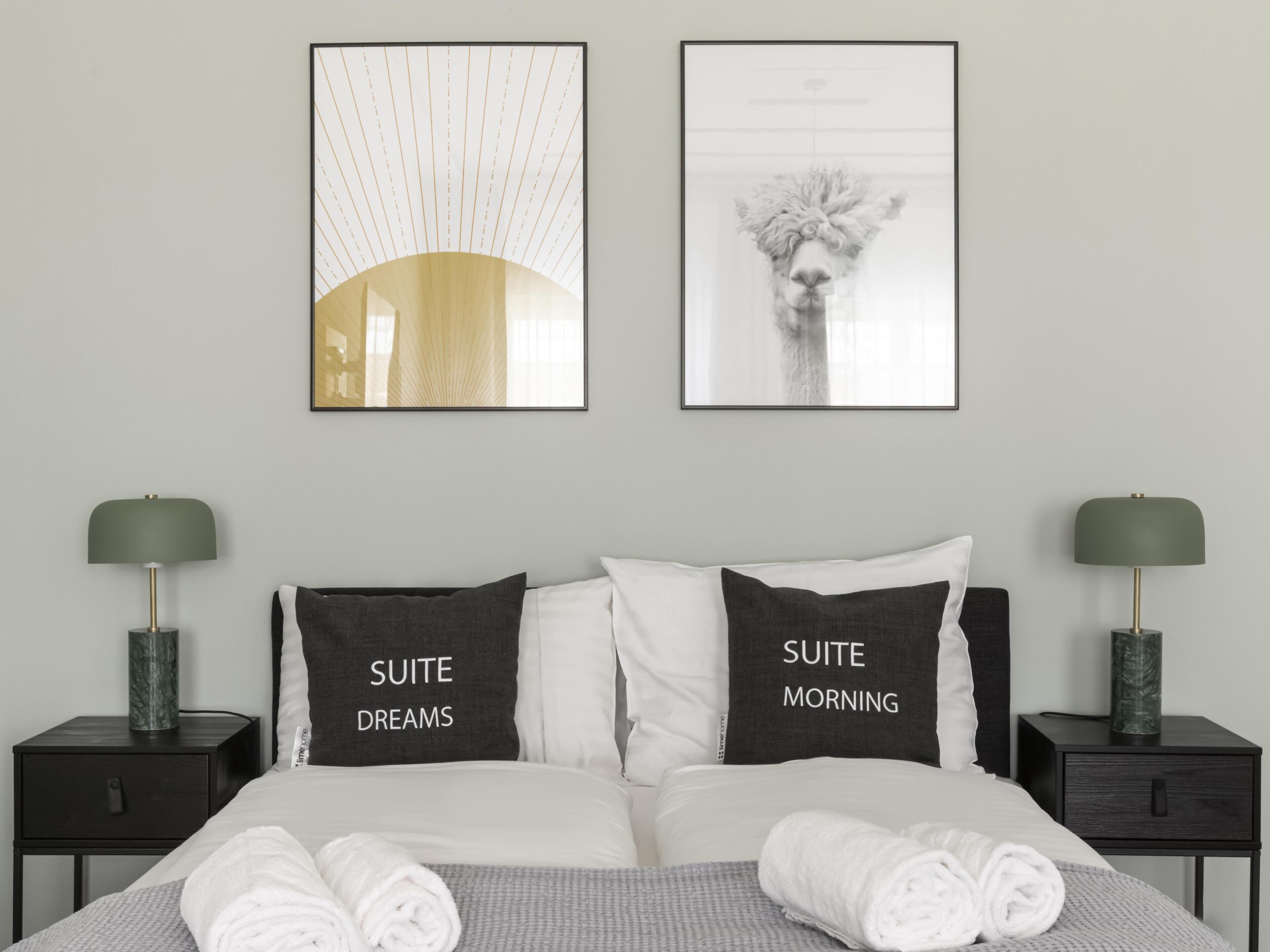 Une chambre d’hôtel avec un lit, deux tables de chevet et deux lampes fixées au mur. Il y a deux oreillers sur le lit, l’un portant l’inscription « RÊVES DE SUITE » et l’autre « MATIN DE SUITE ».