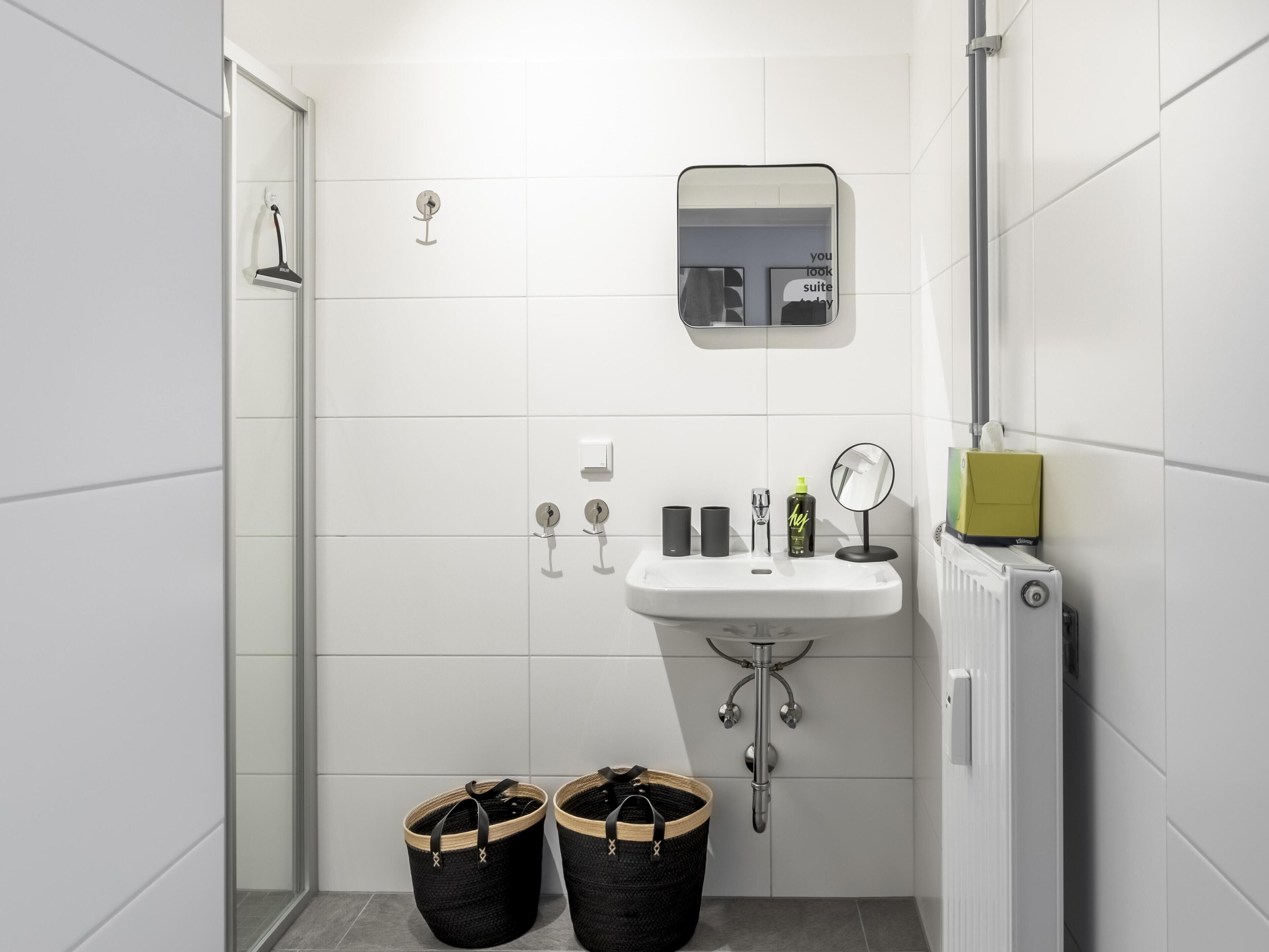 A modern bathroom with a white sink, mirror, and shower. There are two black baskets and a green bottle on the sink.