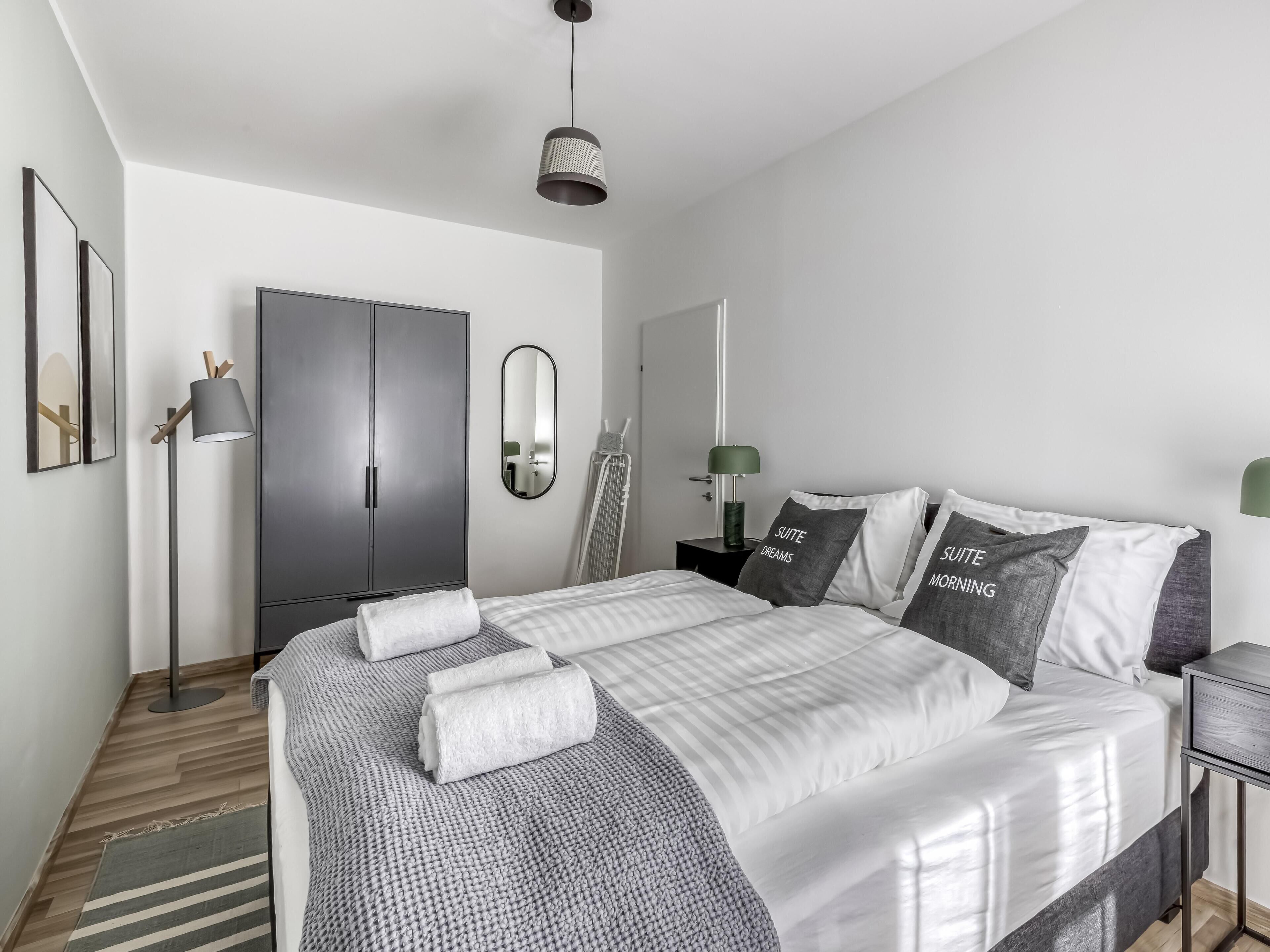 A modern bedroom with a large bed, a wardrobe, a mirror, and a floor lamp.
