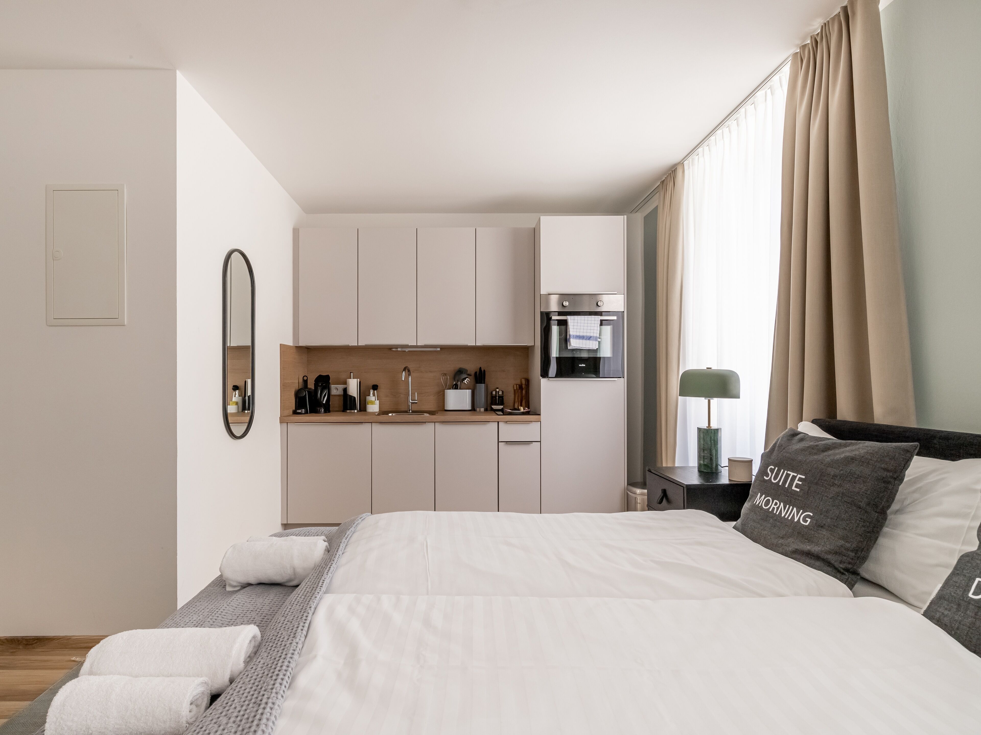 Una camera d'albergo moderna con un letto, un angolo cottura, uno specchio e una finestra con tende.