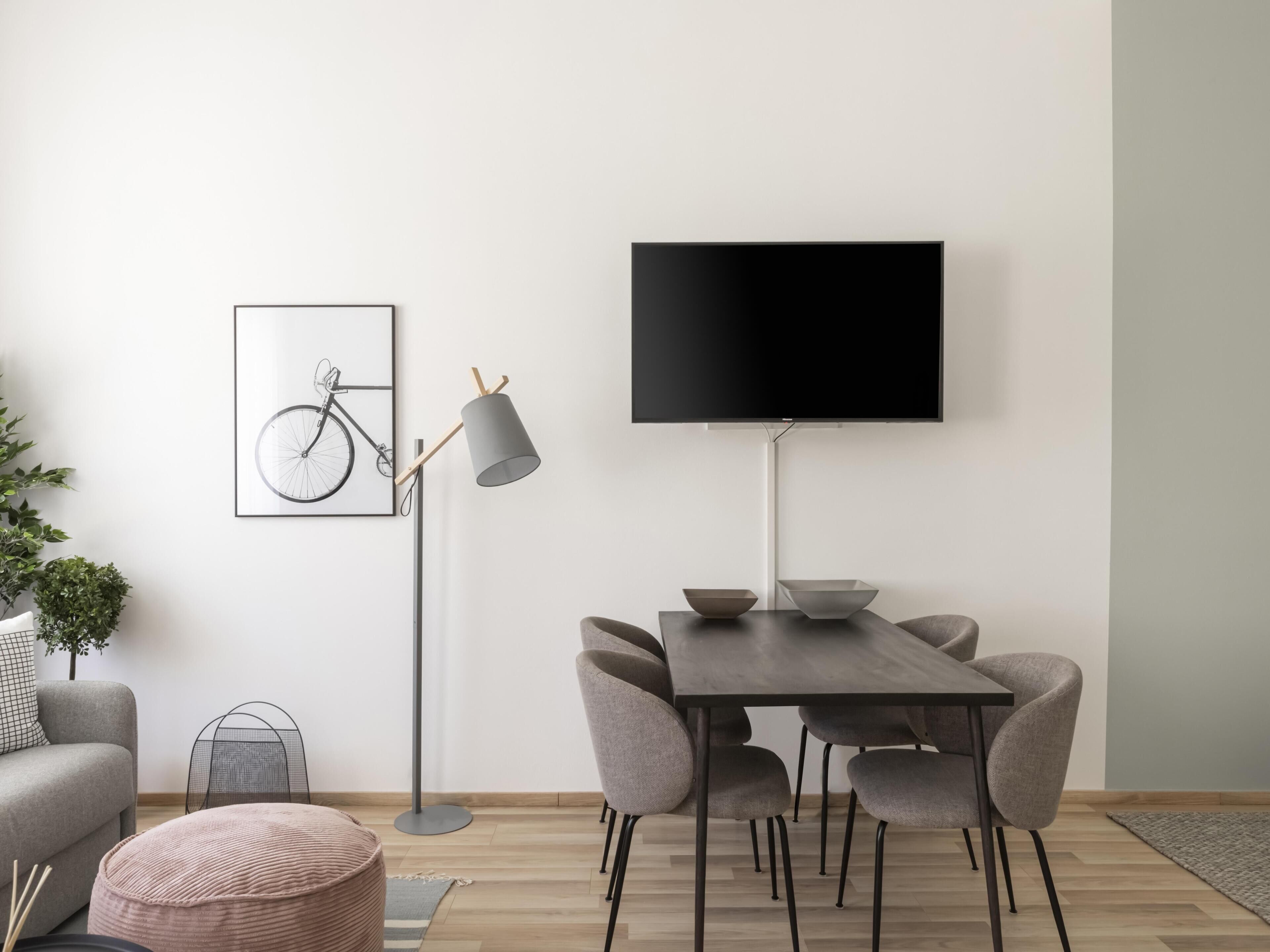 Un soggiorno moderno con un divano grigio, un pouf rotondo, un tavolo da pranzo con sedie grigie, una lampada da terra, un quadro con un'immagine di una bicicletta e un televisore a parete.
