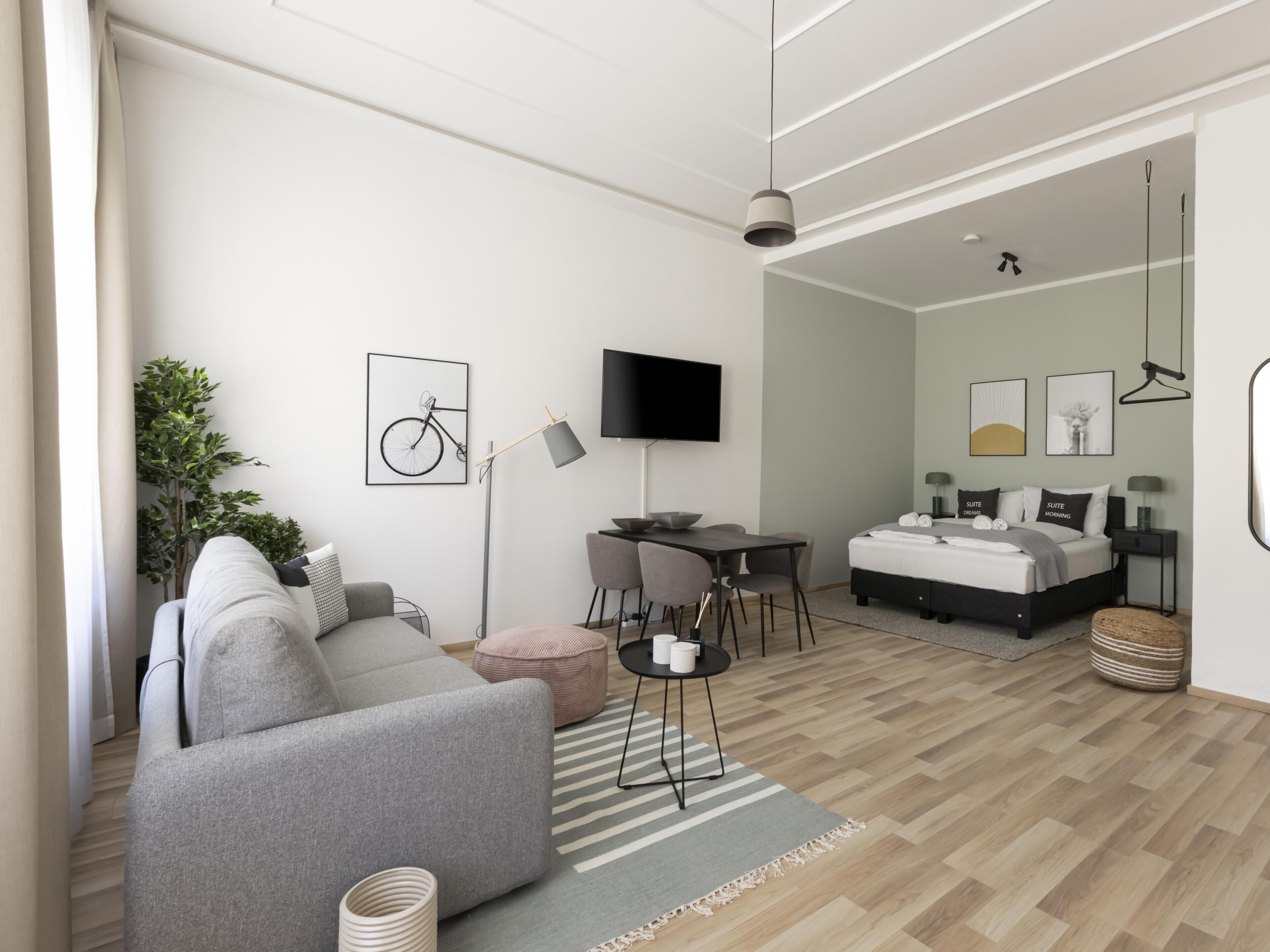 Un salon moderne et bien éclairé, avec un canapé gris, un coin repas comprenant une table et des chaises, un téléviseur à écran plat fixé au mur et un lit avec une tête de lit.