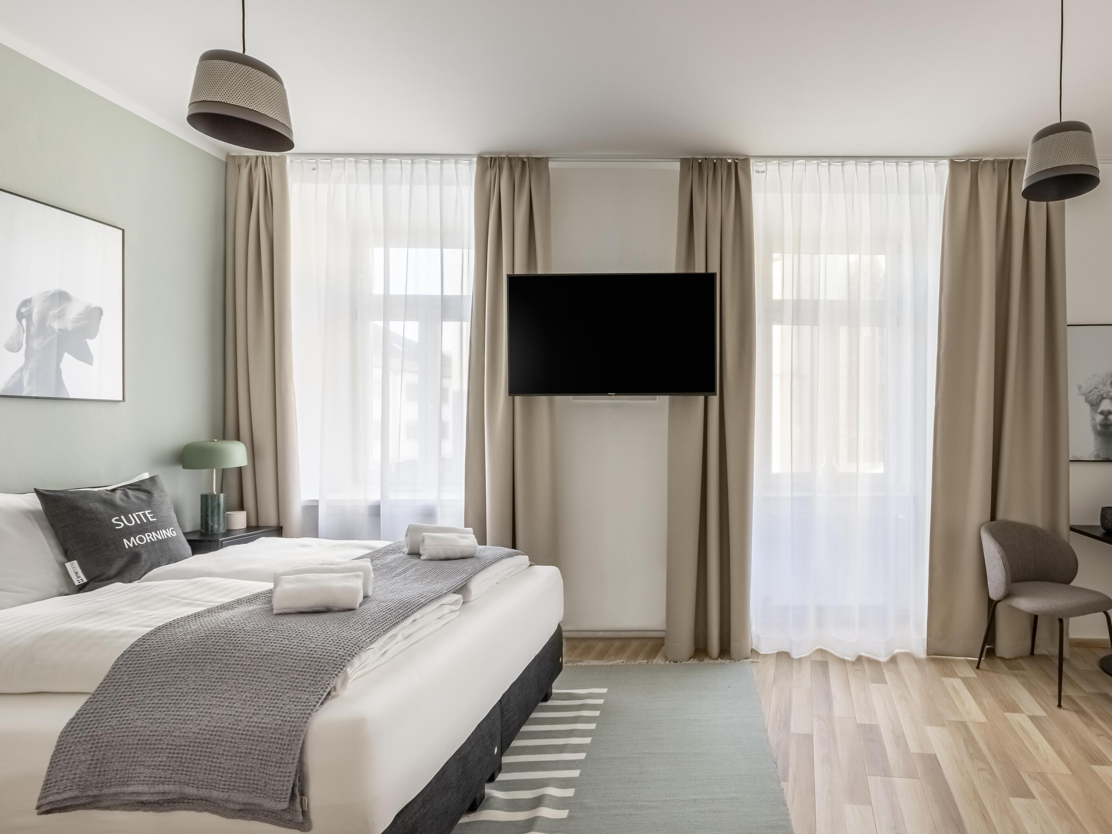 A modern hotel room with a large bed, a flat-screen TV mounted on the wall, and a view of the city through sheer curtains.