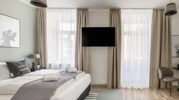A modern hotel room with a large bed, a flat-screen TV mounted on the wall, and a view of the city through sheer curtains.