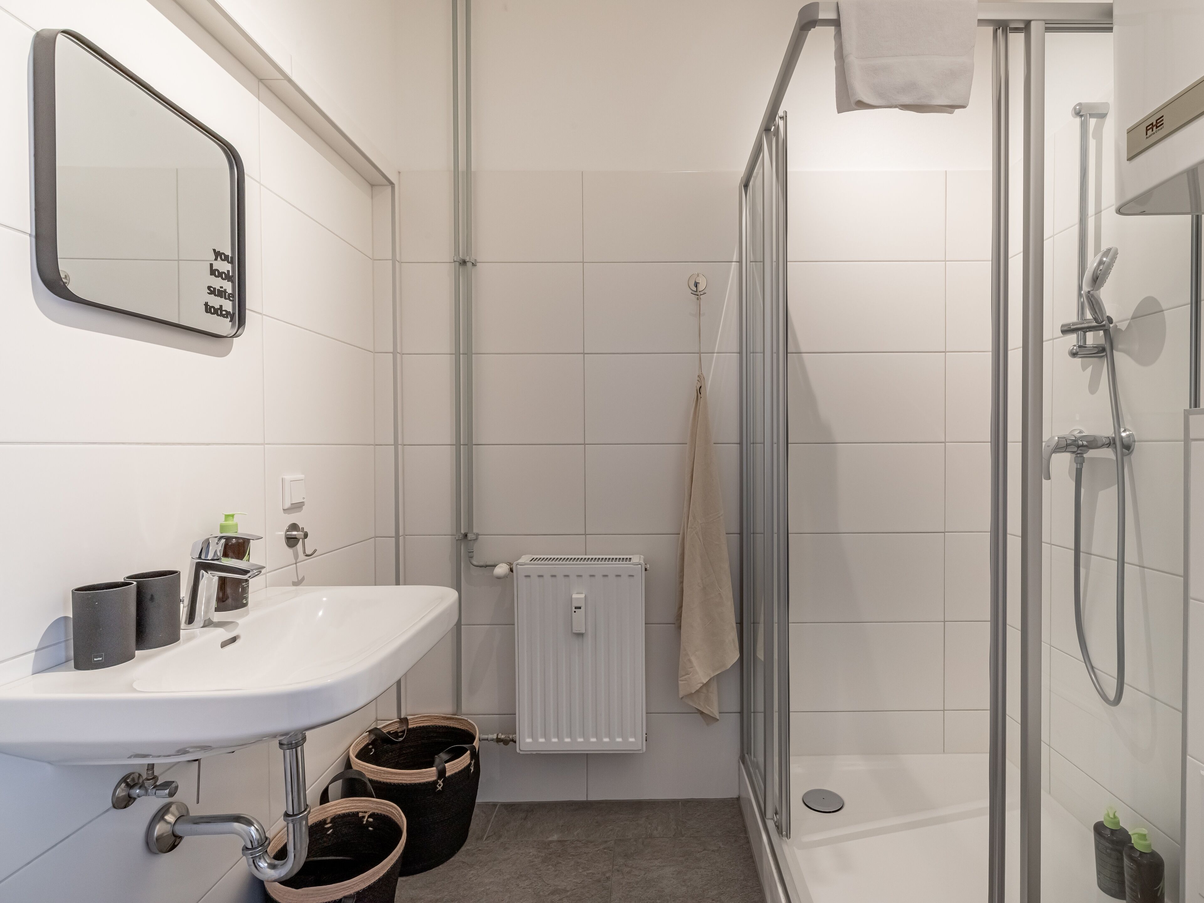 Une salle de bain avec un lavabo blanc, un miroir et une douche. Il y a deux paniers sous le lavabo et une serviette accrochée à la porte de la douche.
