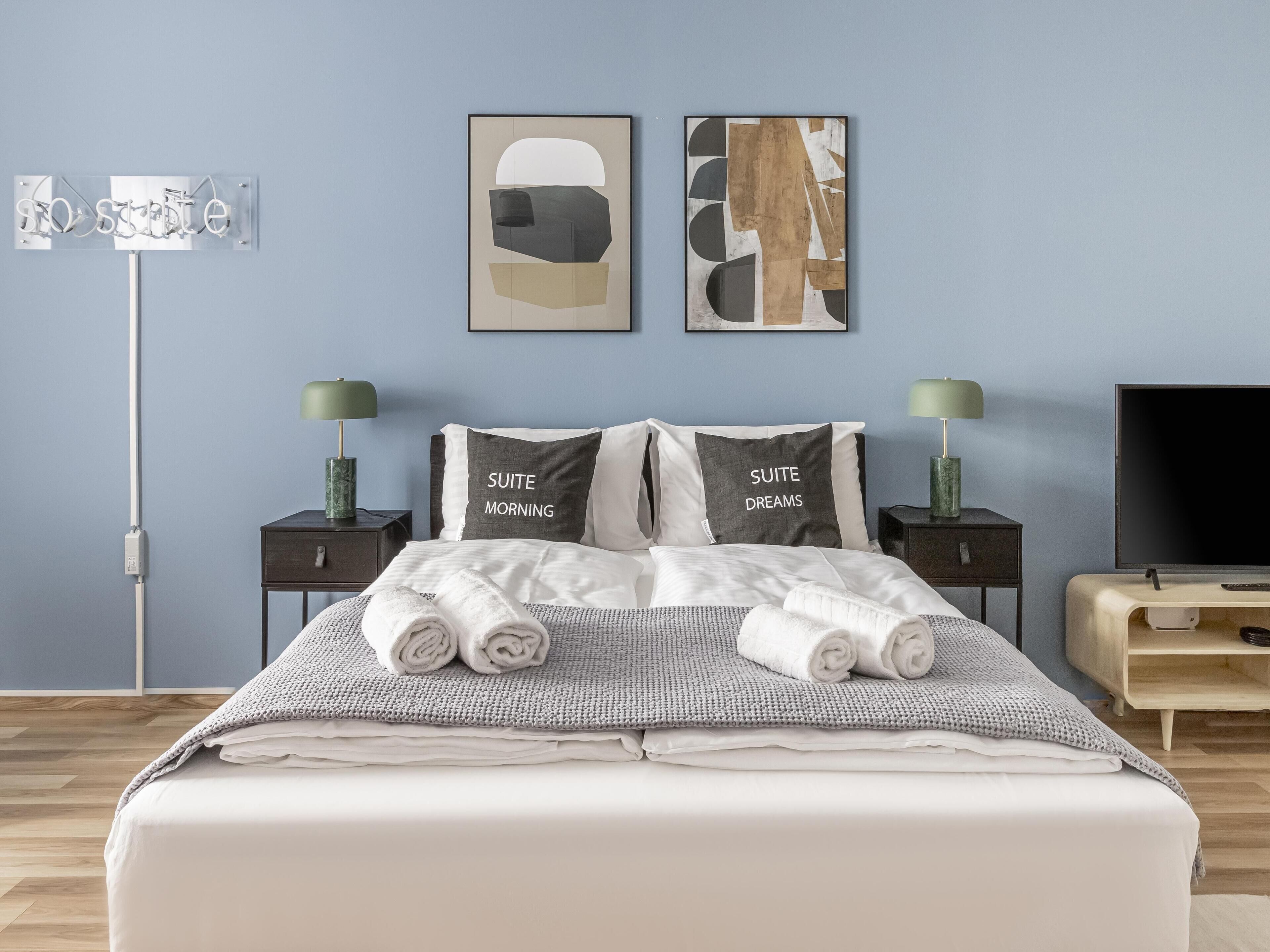 A neatly made bed with two pillows, a bedside table with a lamp, a TV, and artwork on the wall.