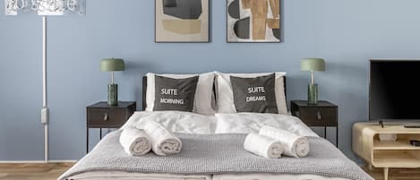 A neatly made bed with two pillows, a bedside table with a lamp, a TV, and artwork on the wall.
