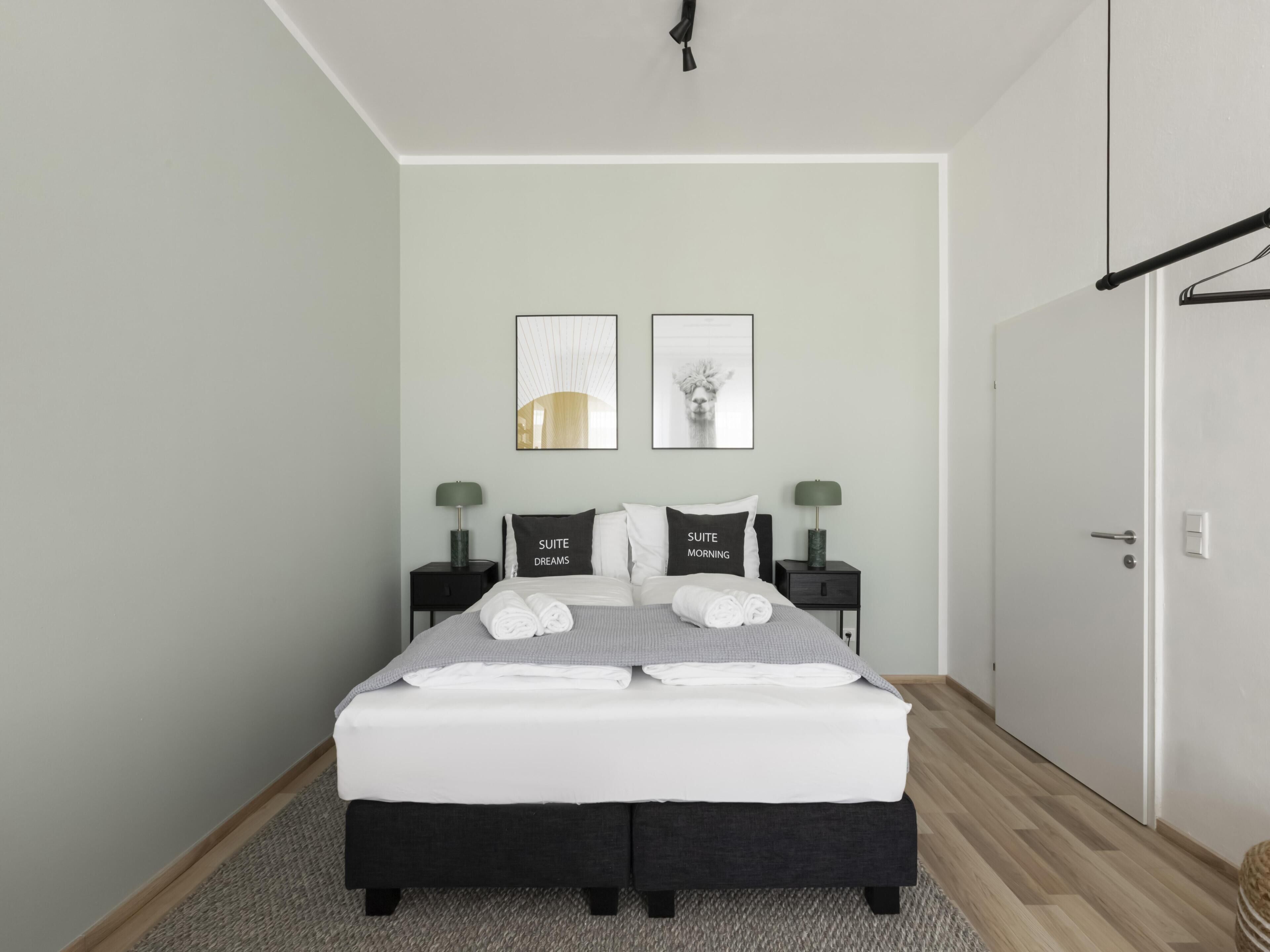 Ein ordentlich eingerichtetes Schlafzimmer mit einem Bett, zwei Nachttischen, einer Tür und zwei gerahmten Bildern an der Wand.