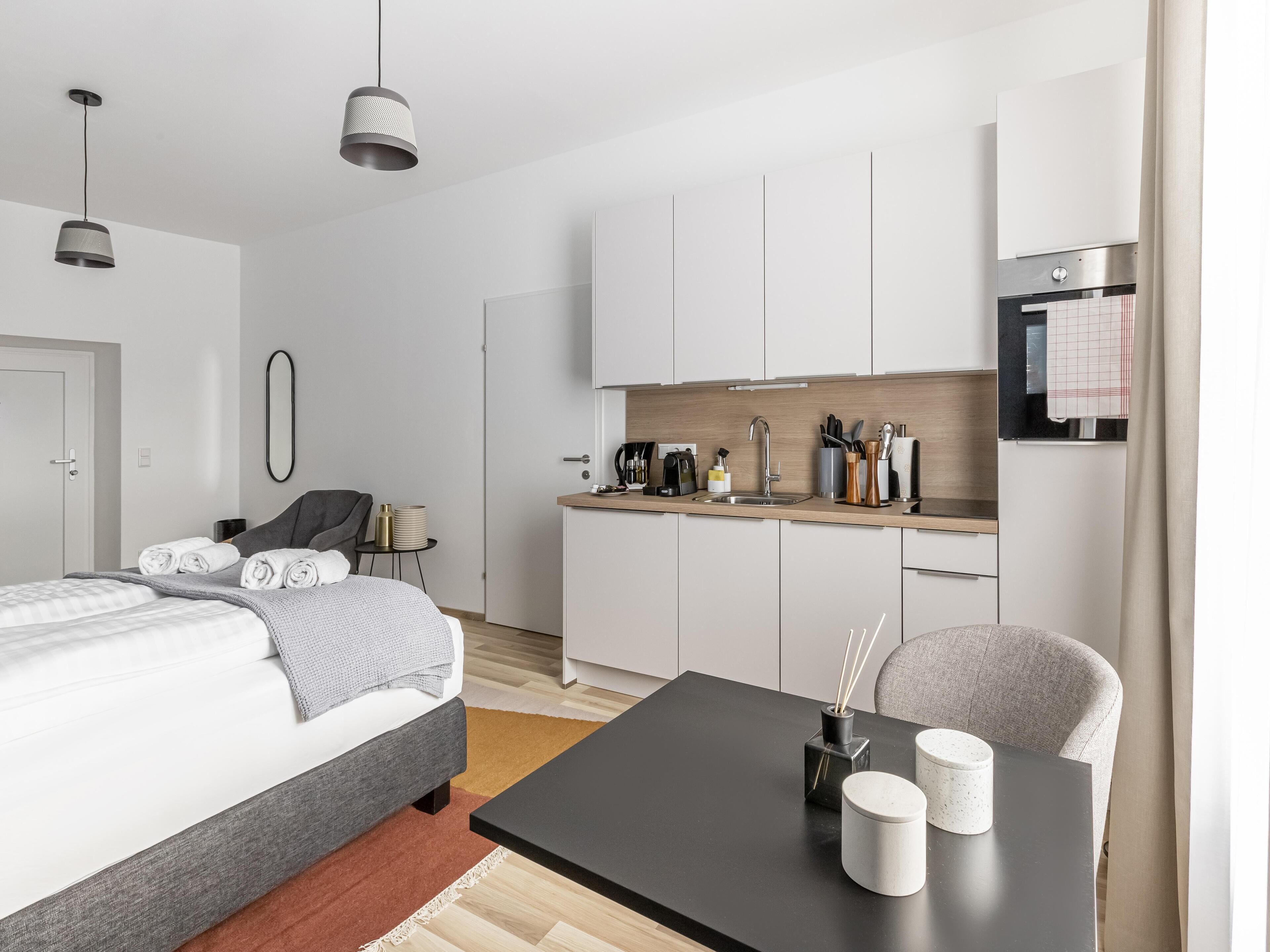A modern, minimalist hotel room with a bed, bedside table, and a small kitchenette.