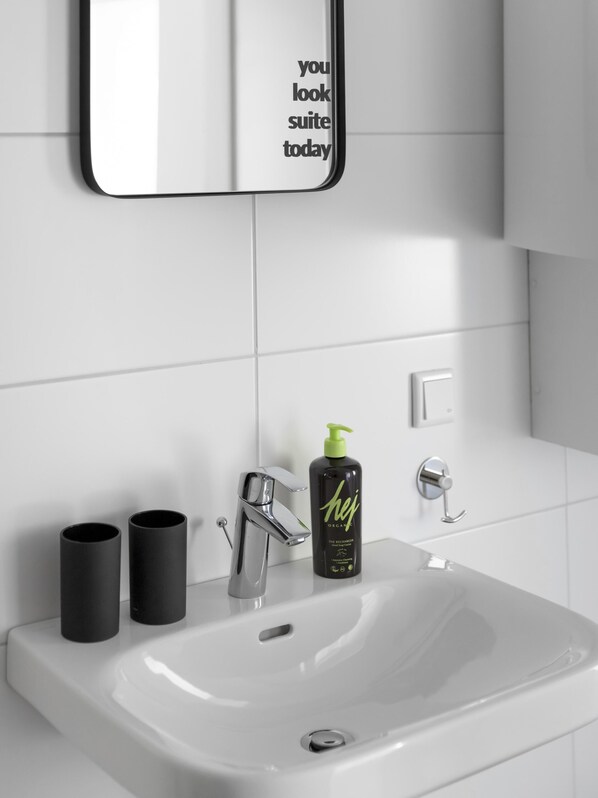 A modern bathroom with a white sink, chrome faucet, and a mirror with motivational text. There are two black cylindrical containers and a bottle of organic hair care product on the sink. - Limehome Villach Kirchenplatz (Villach)