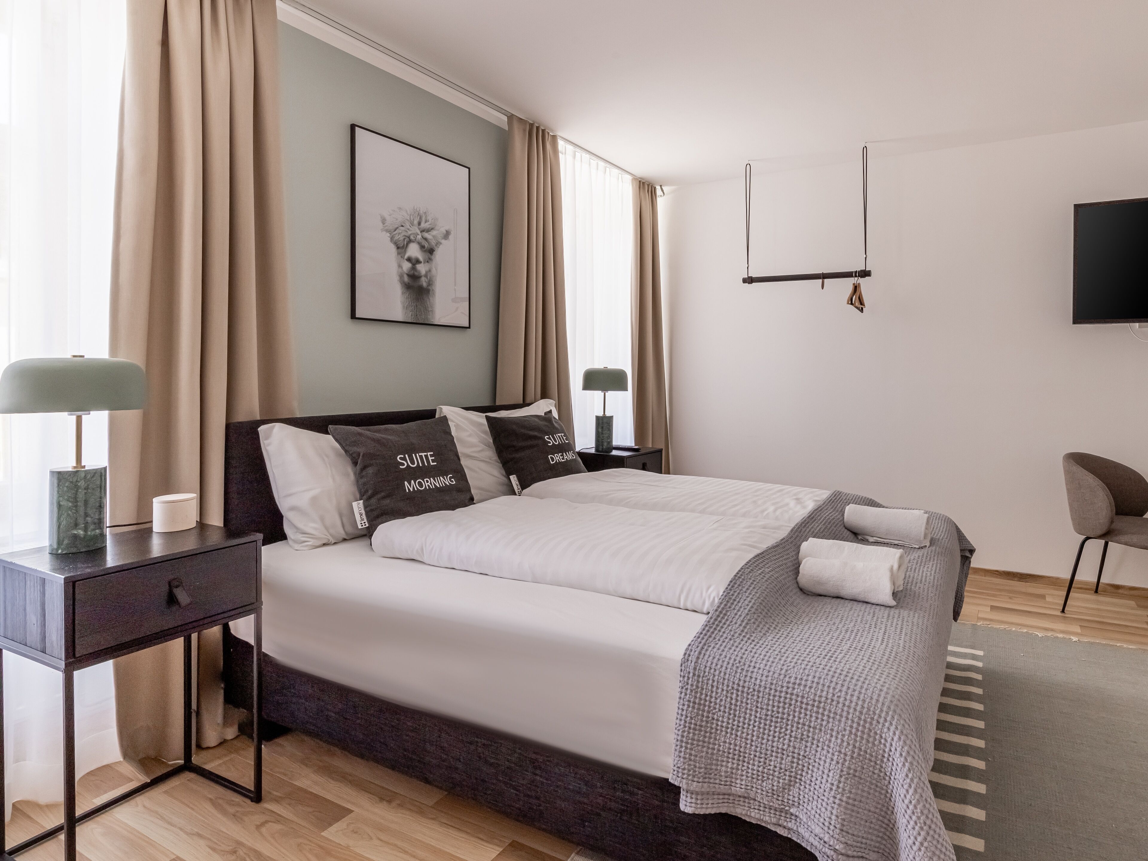 A modern hotel room with a large bed, two bedside tables, a desk, a chair, and a TV on the wall.