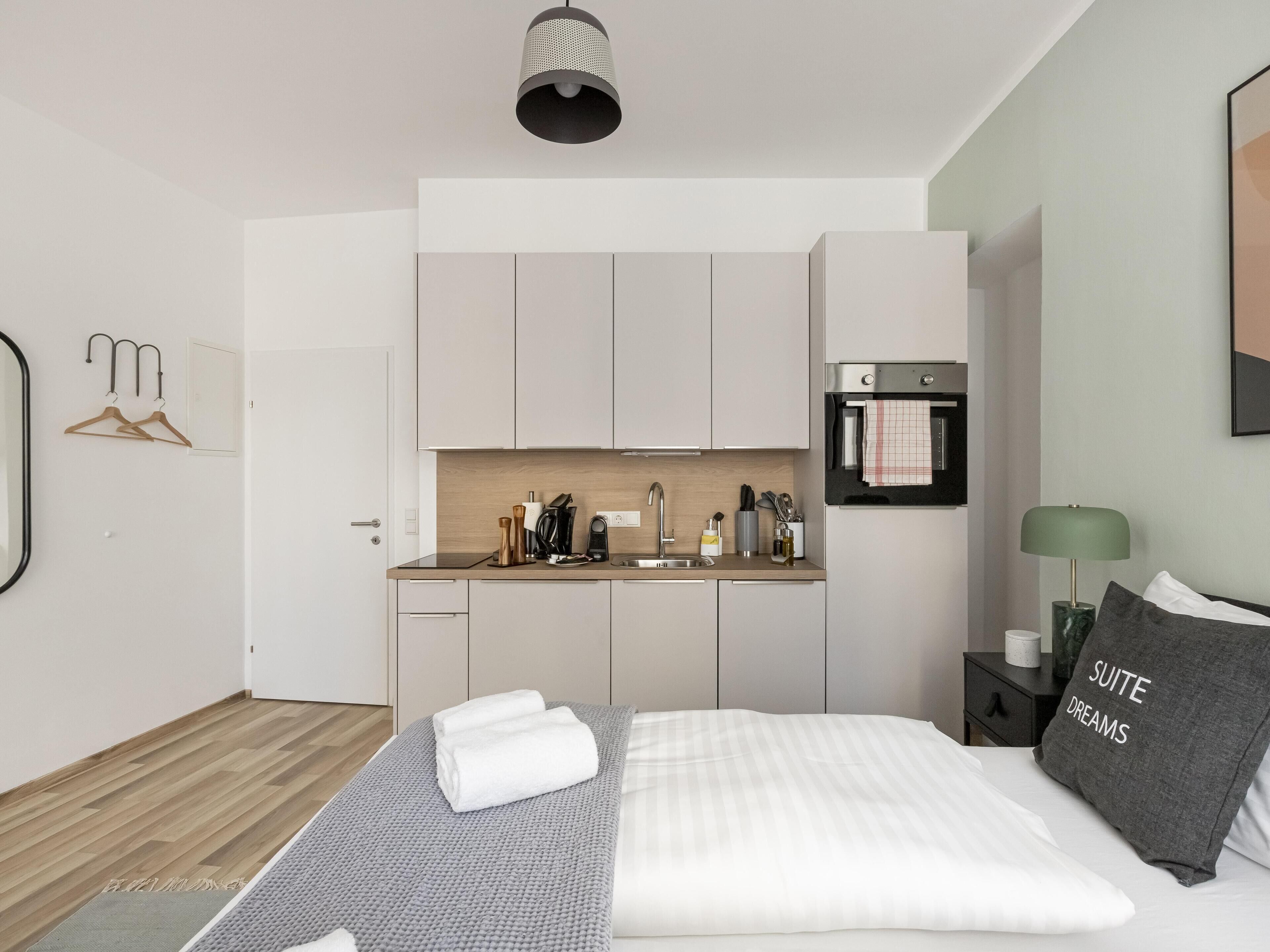 Una habitación de hotel moderna con una cama, una mesita de noche con lámpara, una kitchenette pequeña y la puerta del baño.