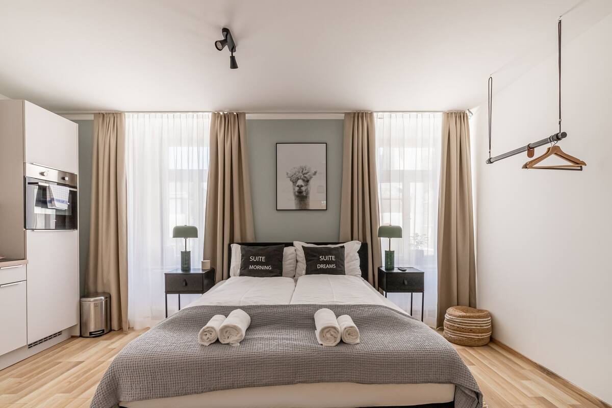 A modern hotel room with a large bed, two bedside tables, a hanging light fixture, and a view of the city through large windows.
