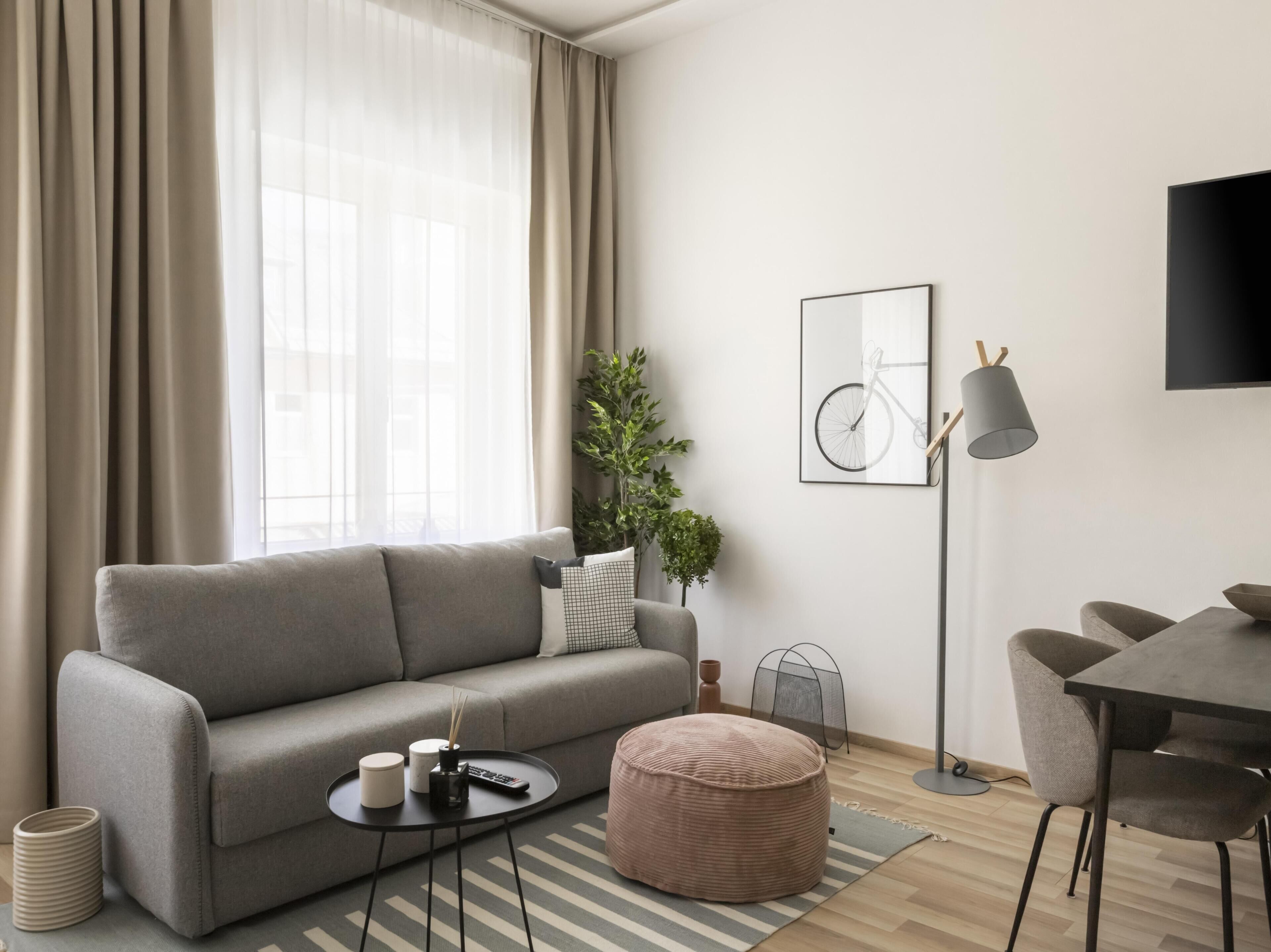A modern living room with a grey sofa, a round ottoman, a small side table with a remote control, a framed bicycle artwork, and a floor lamp.