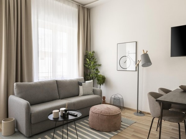 Una sala de estar moderna con un sofá gris, un reposapiés redondo, una pequeña mesita de noche con un control remoto, una obra de arte con una bicicleta enmarcada y una lámpara de pie.