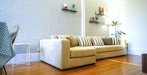 A living room with a beige sofa, a small side table, a white brick wall, and wooden flooring.