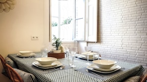 A dining table set with plates, bowls, glasses, and cutlery, with a potted plant and a window in the background.